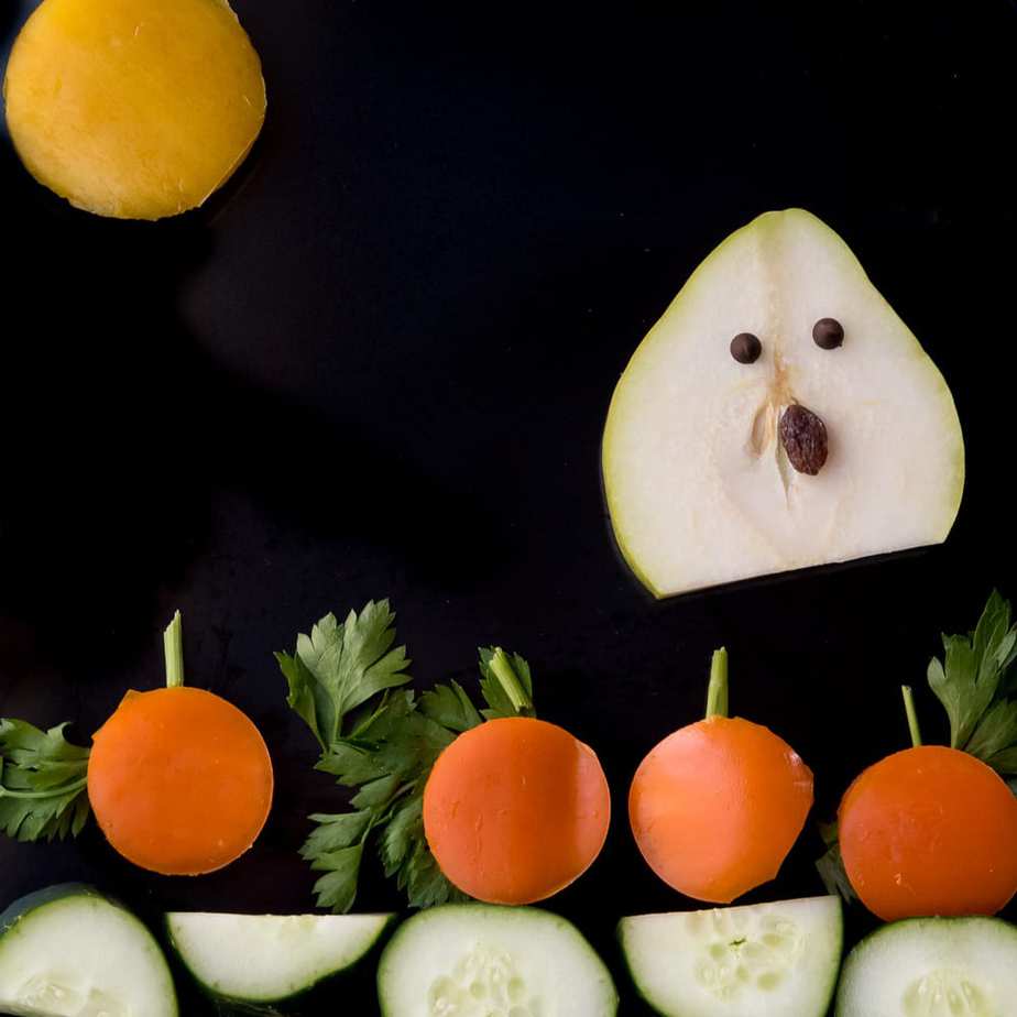 Healthy Paleo halloween snack for kids of a pear sliced into a ghost with raisins for his face, bell peppers shaped into pumpkins and a moon, and parsley for pumpkin stems, and cucumbers for the ground.