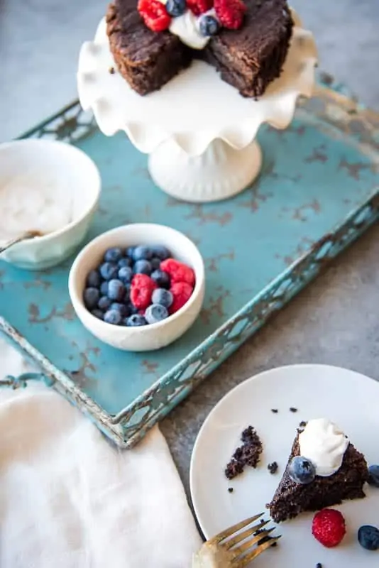 A scene of a chocolate cake on a white stand with fresh berries and coconut whipped cream on a turquoise tray