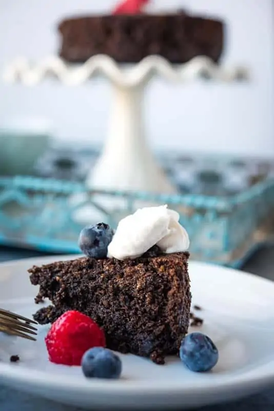 A scene with a closeup of a slice chocolate cake and the rest of the cake in the background