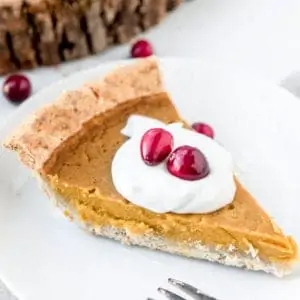 A side view of a slice of pumpkin pie with a topping of whipped cream and fresh cranberries