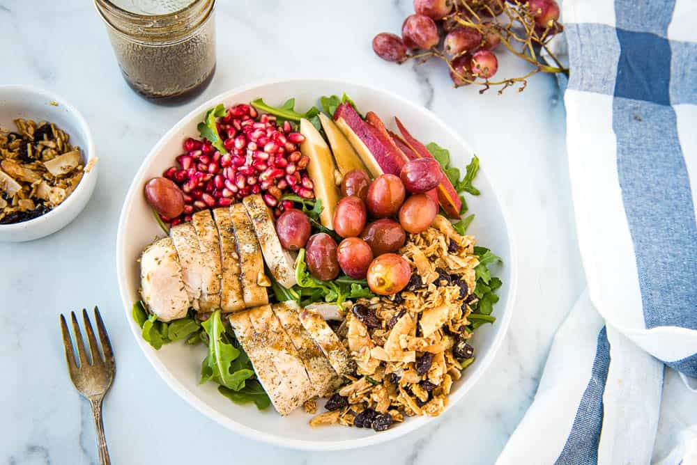 Overhead view of completed pear salad with pomogranete arils, red pears, grapes, cinnamon raisin tigernut granola, sliced baked chicken breast on a bed of arugula in a white bowel.