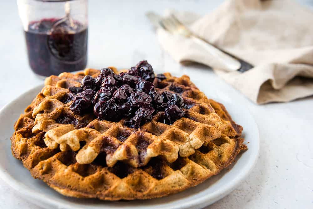 Close up view of two tigernut waffles with blueberry syrup stacked on a white plate with a jar of blueberry syrup and a fork on a napkin in the background.