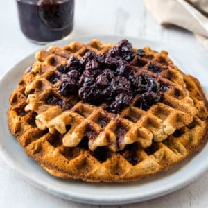 Close up view of two tigernut waffles stacked on a white plate with a jar of blueberry syrup and a napkin in the background.