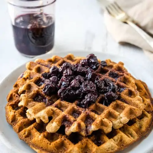 Eye level angle of two tigernut waffles stacked on a white plate with a jar of blueberry syrup and a fork on a napkin in the background.