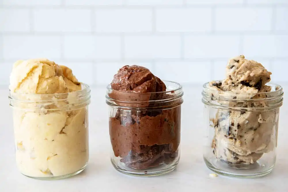 A side view of three single-portion sized mason jars, filled with scoops of AIP Ice Cream. Each jar contains a single flavor; on the right is a jar of Chocolate Chip Ice Cream, in the middle a jar of Cherry Chocolate Ice Cream and at the left a jar of Pina Colada Ice Cream.
