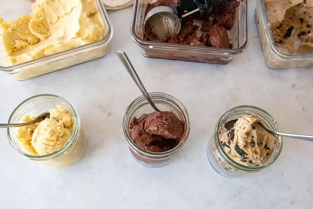 An overhead view of three different homemade AIP Ice Creams. At the front of the picture there are three single portion sized mason jars, filled with scoops of Ice Cream, and behind each, at the top of the picture, is the glass containers of the corresponding ice cream; at the right is the Chocolate Chip flavor, in the middle the Cherry Chocolate flavor and at the left the Pina Colada flavor.