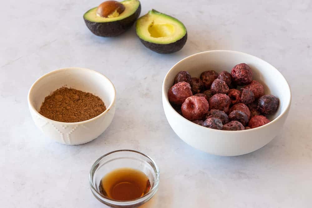 A slightly overhead view of the ingredients for AIP Cherry Chocolate Ice Cream; a breakfast sized white ceramic bowl with frozen cherries to the right, a smaller white ceramic bowl with carob powder to the left, an avocado cut in half, with the seed still inside one of the halves, placed at the top of the picture, and a small glass bowl of maple syrup placed in the front.