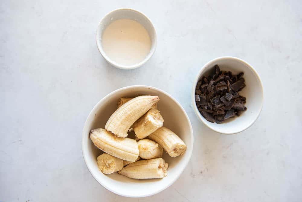 A overhead view of the ingredients for AIP Chocolate Chip Ice Cream; a breakfast sized, white, ceramic bowled with frozen bananas placed at the bottom of the picture, a smaller bowl with carob chips placed a little higher up on the picture and slightly to the right, and a small bowl of milk placed at the top of the picture, slightly to the left.